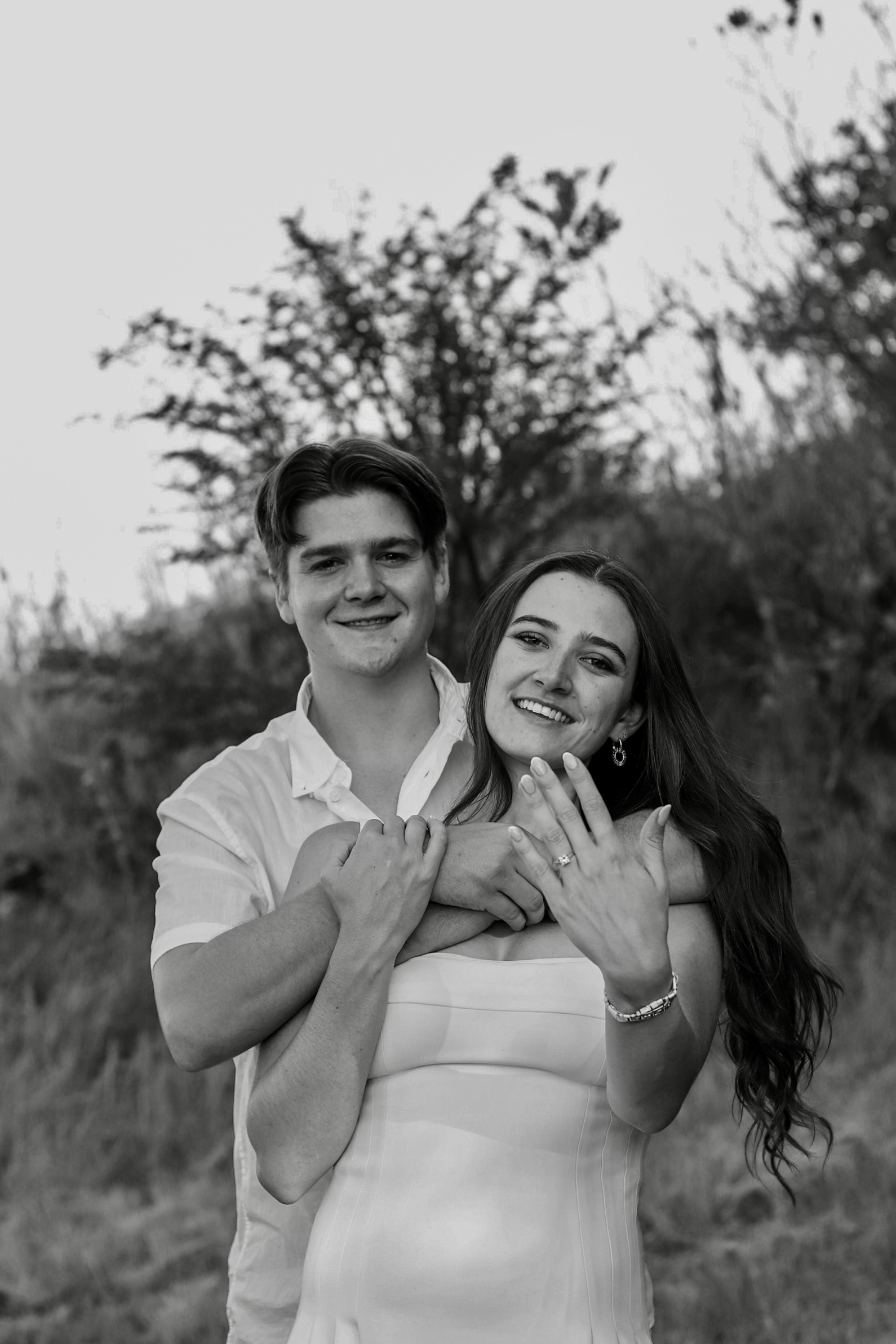 Engagement portrait of a couple embracing outdoors, her hand showing her engagement ring.
