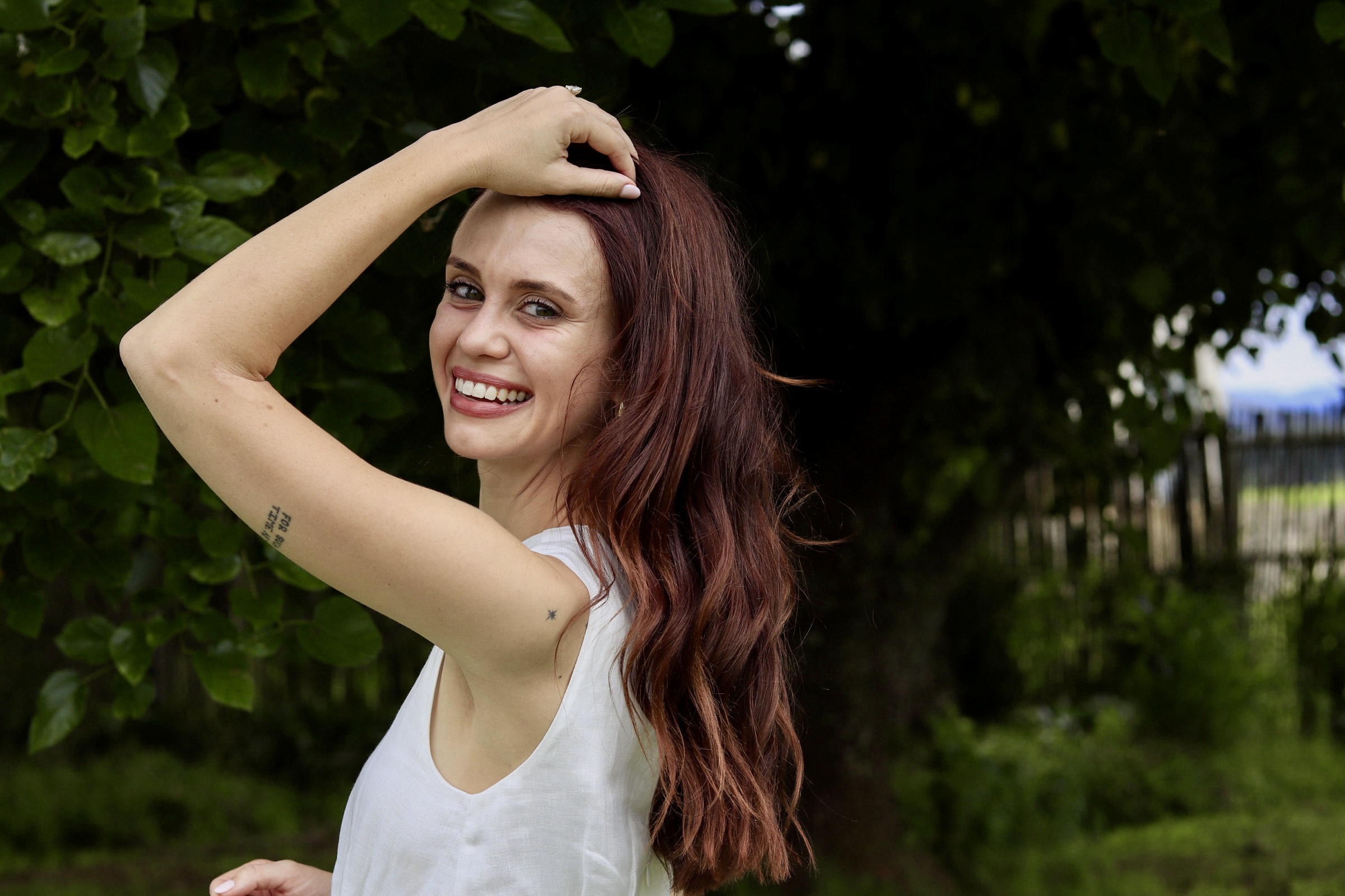 A laughing portrait of a woman with auburn hair, arm raised to her hair.