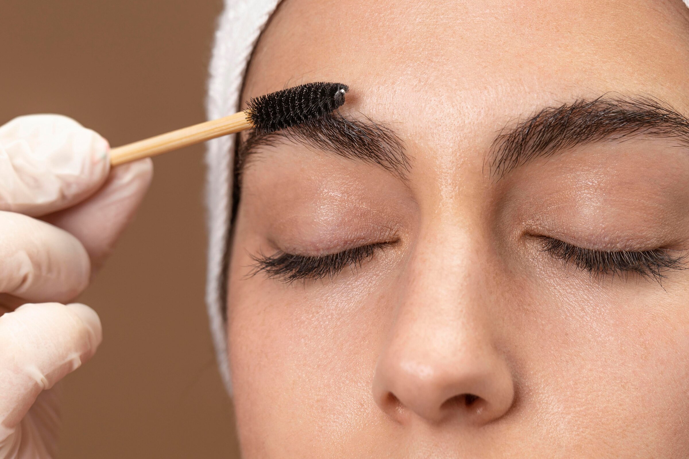 A gloved hand brushing an eyebrow into shape with a small spoolie brush, eyes closed.